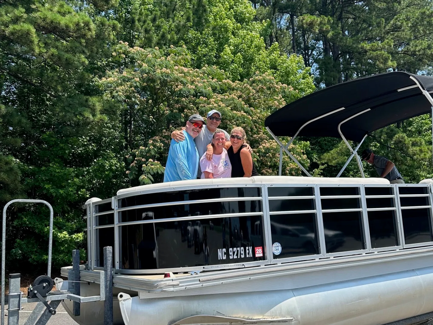 Four people standing on a pontoon boat parked on a trailer, surrounded by tall trees.