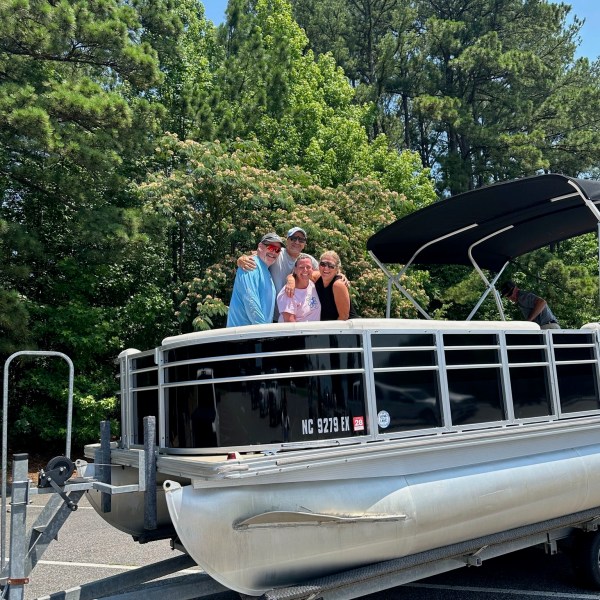 Four people standing on a pontoon boat parked on a trailer, surrounded by tall trees.