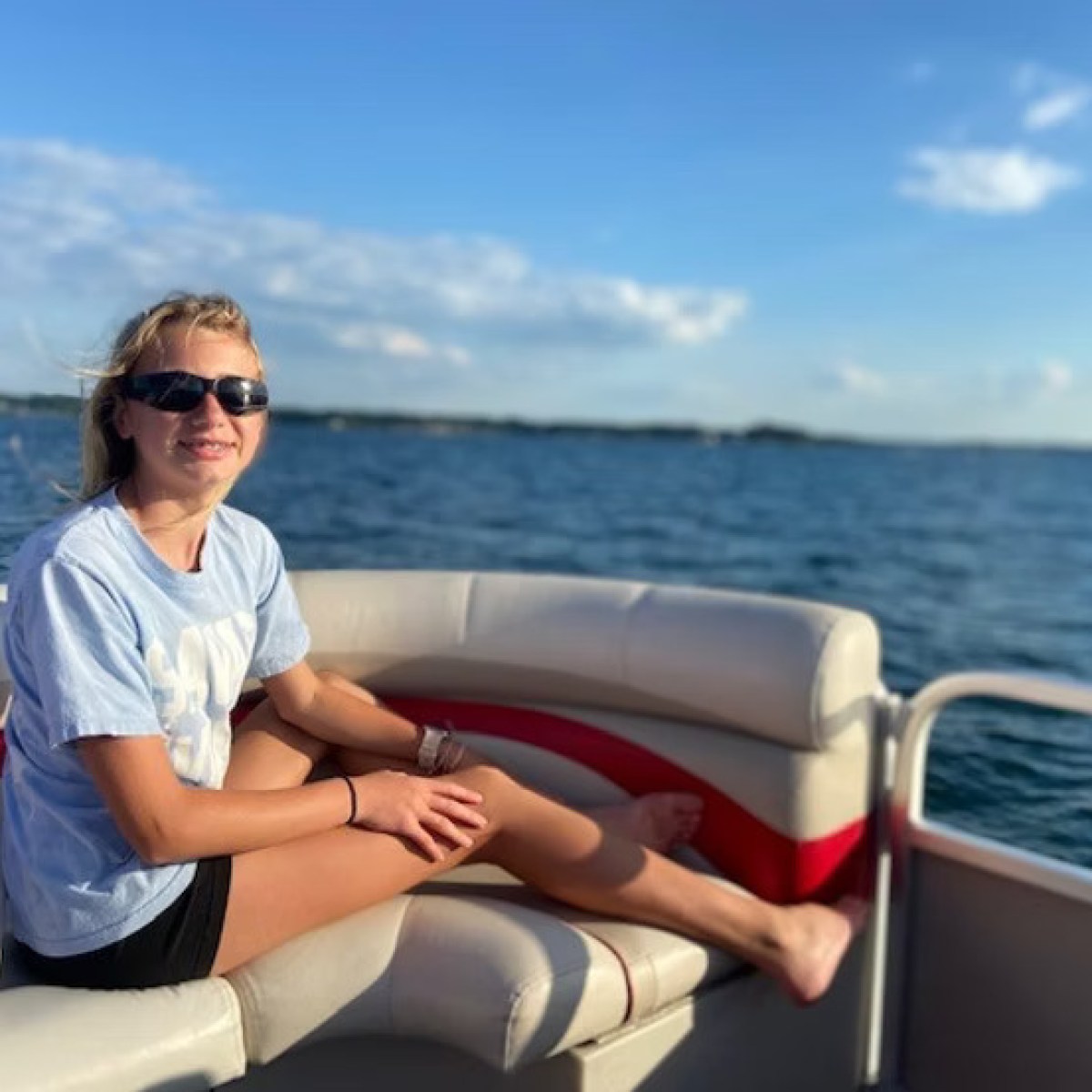 Person wearing sunglasses relaxing on a boat with a lake and blue sky in the background.