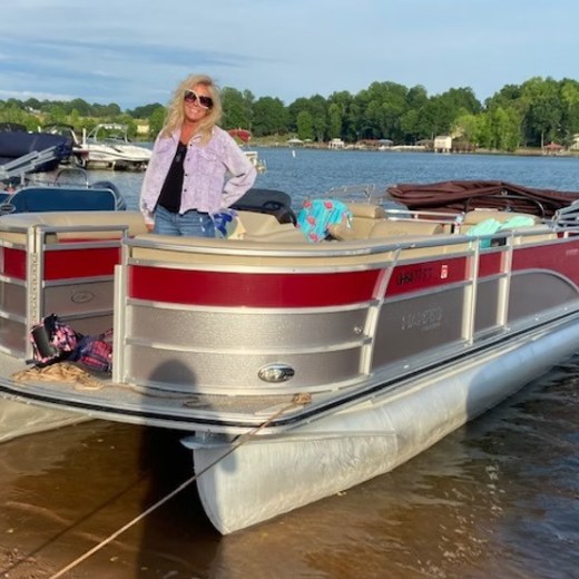 Person standing on a pontoon boat with red trim, docked at a lake.