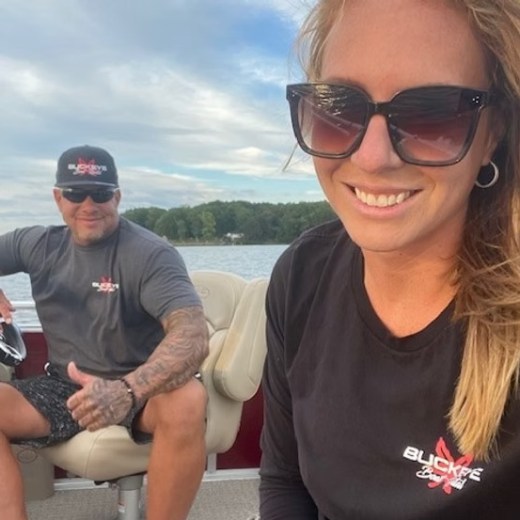 Two people on a boat, wearing sunglasses and smiling, with water and trees in the background.