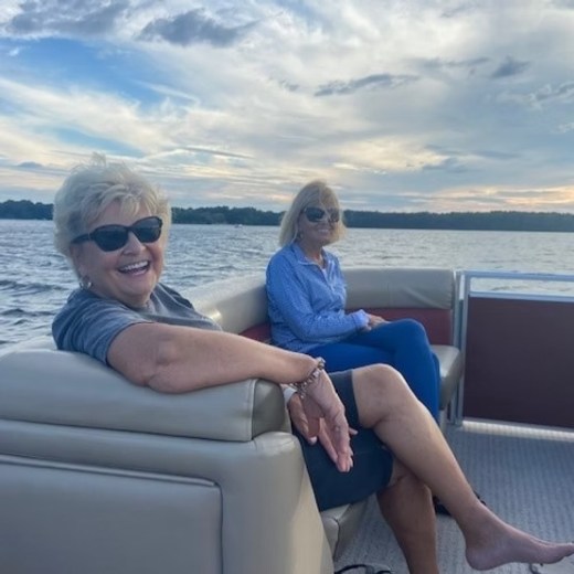 Two women smiling and sitting on a boat with a lake and cloudy sky in the background.