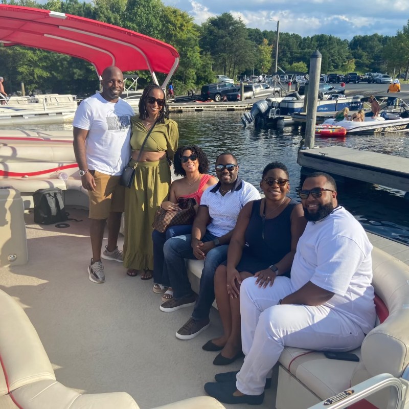 Six people smiling on a boat at a marina with trees in the background.