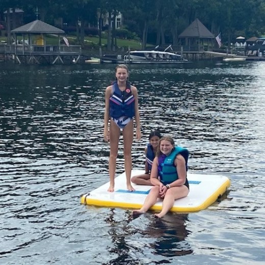 Three people on a floating mat in a lake with boats and pavilions in the background.