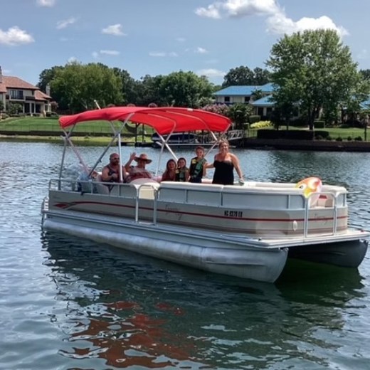 People on a pontoon boat with a red canopy on a lake, surrounded by trees and houses.