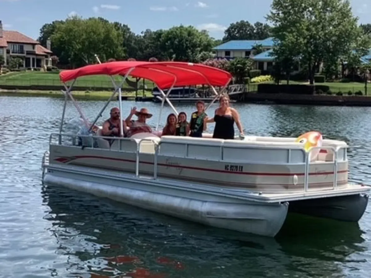Group of people on a pontoon boat with red canopy on a lake, houses and trees in the background.