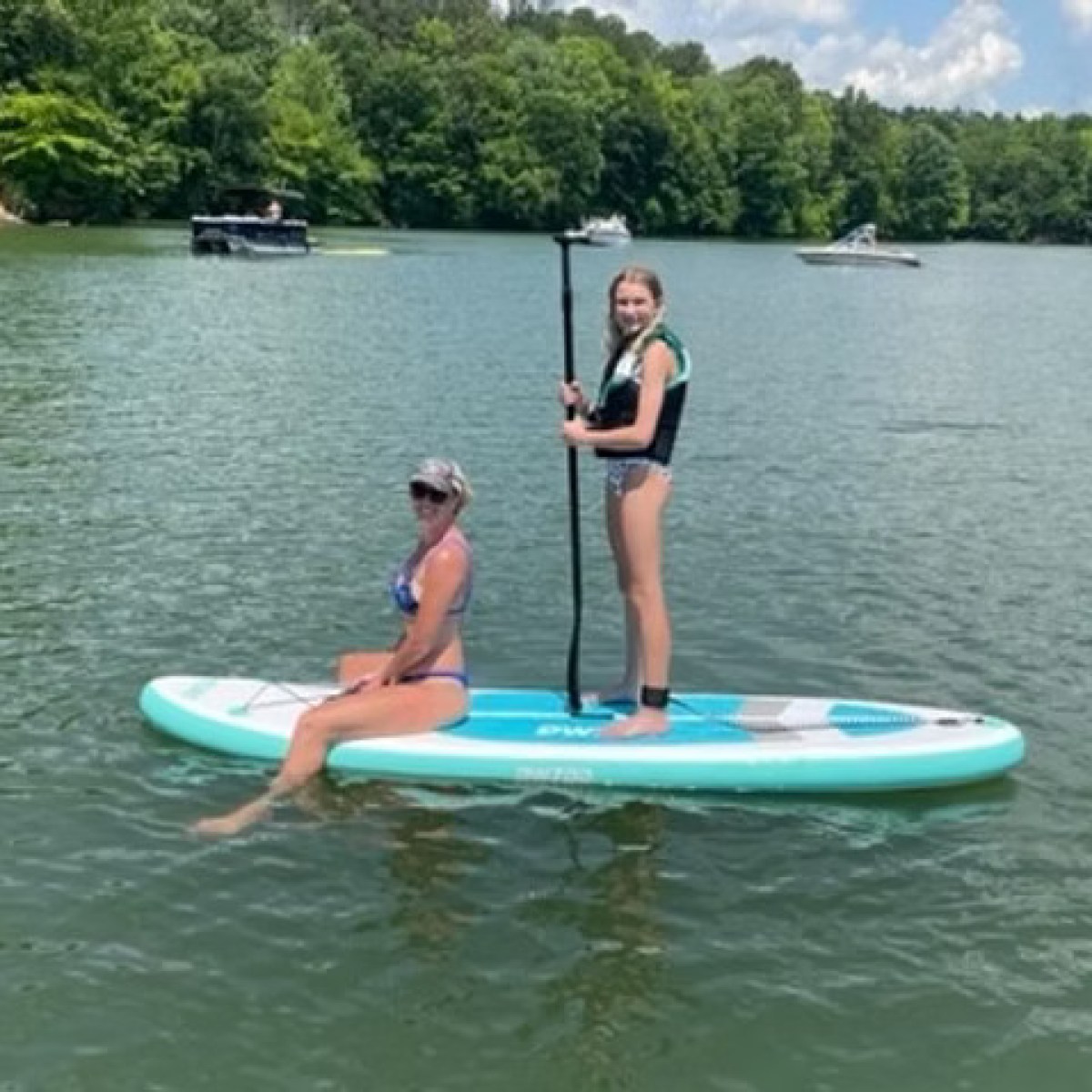 Two people on a paddleboard in a lake; one seated, one standing with a paddle.