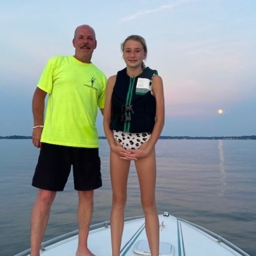 Two people stand on a boat at sunset; one wears a bright shirt, the other a life jacket.