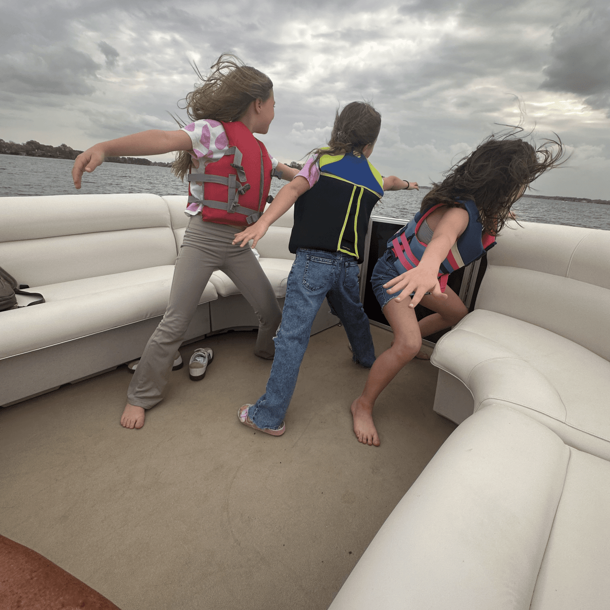 Three kids in life jackets playing on a boat with cloudy skies.