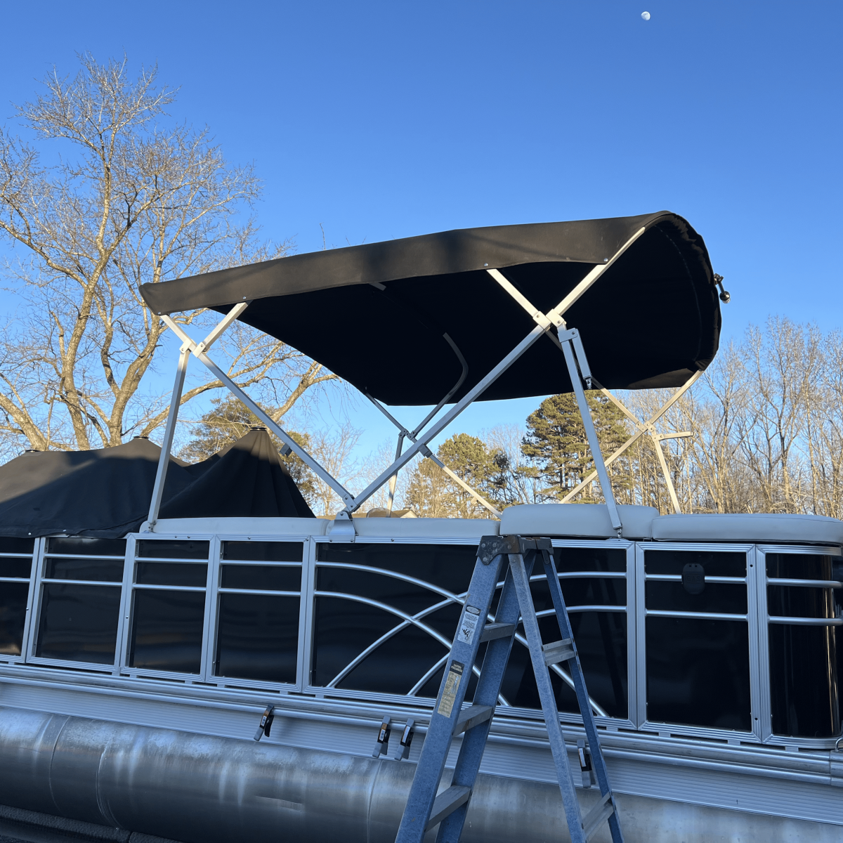 Pontoon boat with a canopy, parked on land near trees and a ladder.